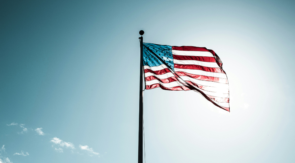 The American flag flying from a flag pole against a blue sky, the sun is shining through the fabric